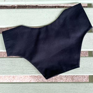 May include: A black face mask made of fabric, with a curved shape designed to cover the nose and mouth. The mask is shown on a light green wooden surface.