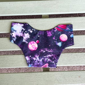 May include: A fabric item with a vibrant galaxy print featuring swirling purples, blues, and pinks. The design includes planets and stars, creating a cosmic theme. The item's shape is curved, suggesting it is a garment.