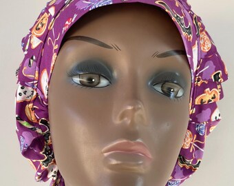 May include: A purple Halloween-themed bouffant scrub cap. The cap features a repeating pattern of pumpkins, candy, bats, and ghosts. The cap has a ruffled edge and is made of a lightweight fabric.