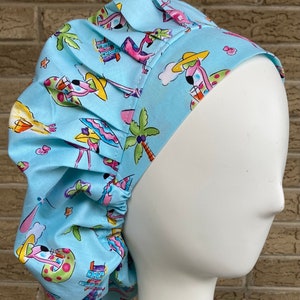 May include: A light blue surgical scrub hat with a pink flamingo and palm tree print. The hat has a gathered elastic band at the back.