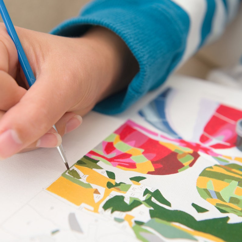 May include: A hand holding a paintbrush over a paint-by-numbers canvas with a colorful abstract design. The canvas features a variety of colors, including red, green, yellow, and blue.