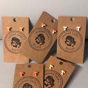 May include: Earrings with a letter "T" design in orange, white, and orange-white combinations. The earrings are displayed on brown cardboard tags with the "Savage Lass Studio" logo. The earrings are small and likely for pierced ears.