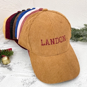 May include: A collection of corduroy baseball caps in various colors, including tan, burgundy, and navy. The tan cap features the name "LANDON" embroidered in red. The caps are arranged in a row, showcasing their different hues and textures.