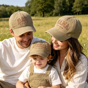 May include: A family wearing matching olive green baseball caps with brown embroidered text. The caps read "DAD EST. 2026", "MAMA EST. 2026", and "ACHILLE EST. 2026". The family is sitting in a grassy field.