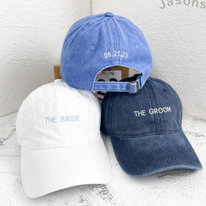 May include: Three baseball caps in blue and white. The white cap has "THE BRIDE" embroidered on the front. The blue cap has "THE GROOM" embroidered on the front. A light blue cap is on top with the date "06.21.25" embroidered on the back.