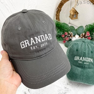 Może przedstawiać: Dwie czapki bejsbolowe, jedna antracytowo-szara i jedna zielona. Szara czapka ma biały haft "GRANDAD EST. 2025". Zielona czapka ma biały haft "GRANDPA EST. 2025". Obie czapki są wykonane z miękkiej tkaniny.