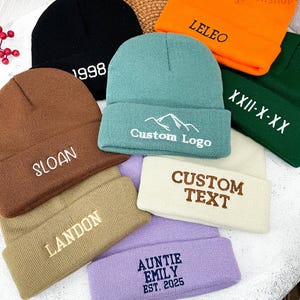 Könnte beinhalten: Eine Auswahl an Mützen in verschiedenen Farben, darunter Schwarz, Braun, Türkis, Orange, Grün, Beige und Lila. Jede Mütze hat gestickte Schriftzüge wie "1998", "SLOAN", "LELEO", "Custom Logo", "XXII-X-XX", "CUSTOM TEXT", "LANDON" und "AUNTIE EMILY EST. 2025".