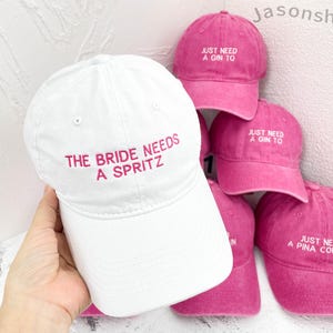 Op de afbeelding: Witte baseballcap met de tekst "THE BRIDE NEEDS A SPRITZ" in roze borduursel. Verschillende roze baseballcaps met de tekst "JUST NEED A GIN TO" en "JUST NEED A PINA COLADA" zijn ook zichtbaar.