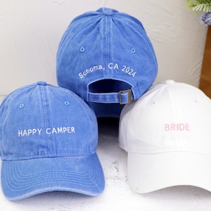 Könnte beinhalten: Drei Baseballkappen aus Denim mit gesticktem Text. Eine Kappe ist blau mit "Happy Camper" in weißer Schrift. Eine weitere Kappe ist blau mit "Sonoma, CA 2024" in weißer Schrift. Die dritte Kappe ist weiß mit "BRIDE" in rosa Schrift.