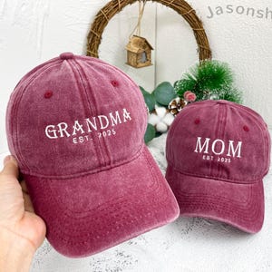 May include: Two red baseball caps with white embroidered text. The first cap says "Grandma Est. 2025" and the second cap says "Mom Est. 2025".