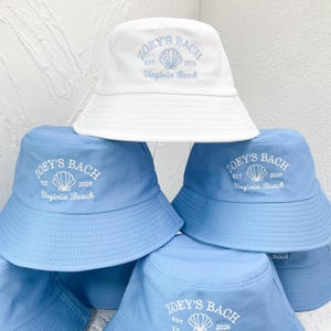 Peut inclure: Un chapeau bob blanc et plusieurs chapeaux bob bleu clair, chacun orné d'une broderie indiquant Zoey's Bach, Est 2026, et Virginia Beach, avec un motif de coquillage central. Les chapeaux ont un bord large et une calotte arrondie.