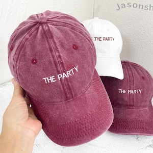 May include: Three baseball caps are displayed. Two are a deep red, and one is white. Each cap features the embroidered text "THE PARTY" in white. The caps are made of a soft, textured material.