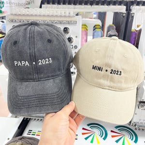 May include: Two baseball caps, one dark gray and one beige, are held up. The dark gray cap has "PAPA • 2023" embroidered in white. The beige cap has "MINI • 2023" embroidered in black. Both caps have a curved brim and adjustable strap.