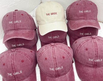 May include: A selection of burgundy baseball caps, some with "THE GIRLS" embroidered in white, and one cream-colored cap with "THE BRIDE". The caps are stacked, displaying their design. The caps are a faded burgundy color.