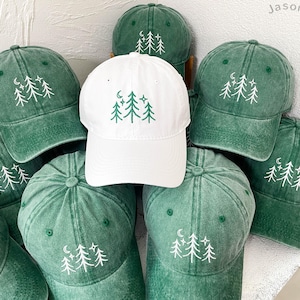 Może przedstawiać: Kolekcja zielonych i białych czapek z daszkiem. Każda czapka ma haftowany wzór drzew, półksiężyca i gwiazd. Zielone czapki mają wyblakły, vintage wygląd.