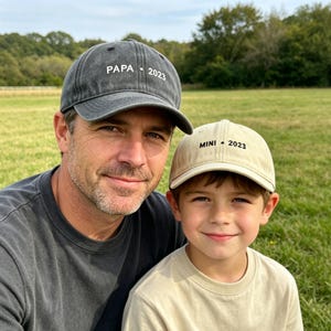 May include: Two baseball caps, one gray and one tan, with the text "PAPA 2023" and "MINI 2023" embroidered on the front. The caps are worn by a man and a child, respectively, in a field.