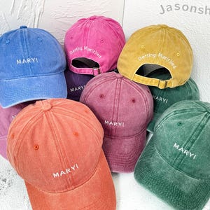 Könnte beinhalten: Ein Set aus neun Baseballkappen in verschiedenen Farben, darunter Pink, Blau, Gelb, Rot, Grün und Braun. Die Kappen haben das Wort "MARY!" in Weiß auf der Vorderseite gestickt. Einige der Kappen haben auch den Spruch "Getting Married" auf der Rückseite gestickt.