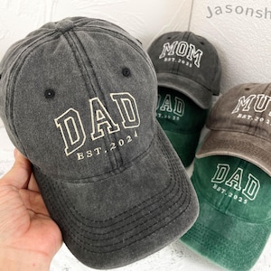 Könnte beinhalten: Vier Baseballkappen aus abgenutztem Stoff mit gesticktem Text. Die Kappen sind schwarz, braun, grün und schwarz. Der Text auf den Kappen lautet "DAD EST. 2024", "MOM EST. 2023", "MUM EST. 2025" und "DAD EST. 2025".