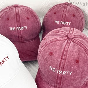 Op de afbeelding: Een verzameling baseballpetten in bordeauxrood en wit. De bordeauxrode petten hebben de witte tekst "THE PARTY". De witte pet heeft ook de rode tekst "THE PARTY". De petten zijn gemaakt van een zacht, getextureerd materiaal.