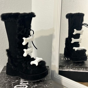 以下が含まれることがあります： 白い骨の形をしたバックルが付いた、黒いフェイクファーの裏地が付いたブーツ。ブーツは厚いプラットフォームソールとレースアップのフロントを持っています。