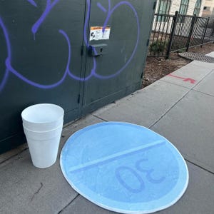 Puede incluir: Una alfombra redonda azul claro con un diseño que se asemeja a una pastilla grande, con el número 30. Junto a ella, una pila de tres vasos de papel blancos descansa sobre una superficie de hormigón. El fondo incluye una caja de servicios pública verde oscuro con graffiti morado.