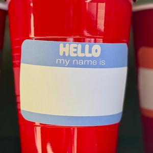 May include: Three red plastic cups with white rims are displayed. Each cup has a label that reads "HELLO my name is" with a blank space for writing a name. The labels have a blue border.