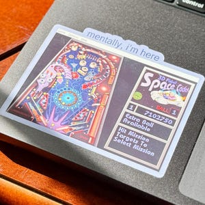 May include: A rectangular sticker featuring a colorful pinball machine design with the text "Space Code." The sticker also includes the text "mentally, I'm here." The sticker is placed on a laptop.