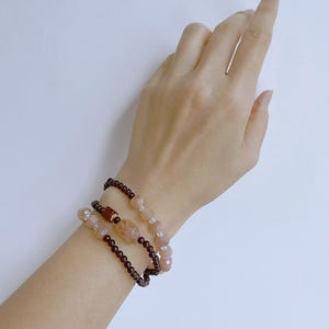 May include: Three beaded bracelets in shades of brown and pink gemstones. The bracelets are stacked on a wrist, with a simple gold ring on a finger. The background is a plain white surface.