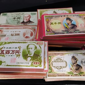 May include: Stacks of Japanese banknotes featuring portraits and intricate designs. The bills display various denominations, including 5,000, 10,000, and 1,000,000 yen notes. The banknotes have a red border and are stacked on top of each other.