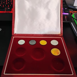 May include: A red velvet coin display case with a white satin interior. The case holds six coins of varying colors, including silver, gray, and gold. The case is open, revealing the coins.