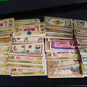 May include: A collection of vintage Japanese Yen banknotes, featuring various denominations. The bills display intricate designs with Japanese text and illustrations, in shades of green, pink, and yellow. The banknotes are stacked in piles, showcasing their aged appearance.