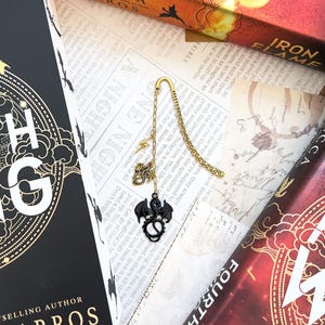 May include: A gold bookmark with a black dragon charm. The bookmark is hanging from a book with the title "A Night in the Iron Flame" by Rebecca Yarros.