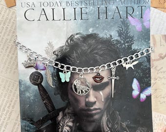 May include: A silver charm bracelet with various charms, including a wolf, a crown, a feather, a butterfly, a dagger, and a lip. The bracelet is on top of a book cover with the title "Quicksilver" and the author's name "Callie Hart".