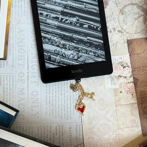 Puede incluir: Un lector electrónico Kindle negro con un charm de marcapáginas dorado y rojo. El lector electrónico muestra una imagen en blanco y negro de una pila de bolígrafos. El texto "ONE NIGHT ONLY" y "A NIGHT OF MAGIC" es visible en el fondo.