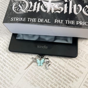 May include: A silver bookmark with a blue butterfly charm, a sword charm, and a feather charm. The bookmark is resting on a book with the text "STRIKE THE DEAL. PAY THE PRICE" and "DREAM OF THE GOOD THINGS TO".