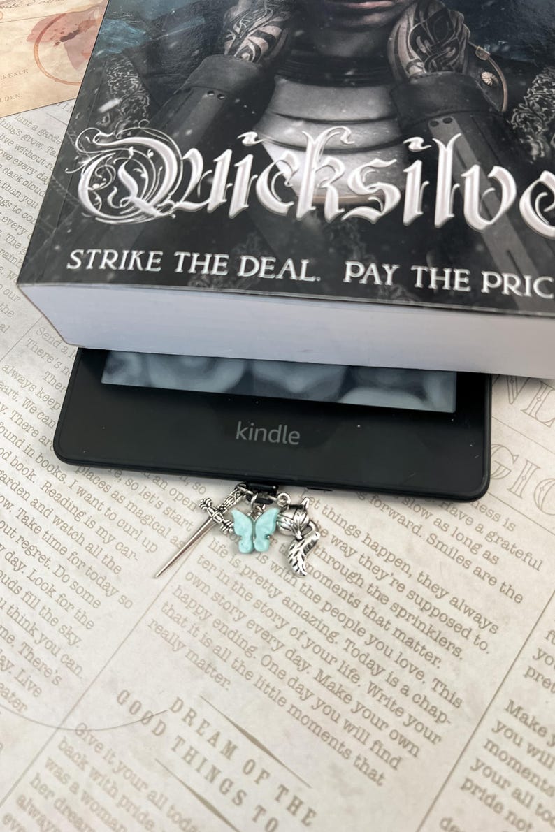 May include: A silver bookmark with a blue butterfly charm, a sword charm, and a feather charm. The bookmark is resting on a book with the text "STRIKE THE DEAL. PAY THE PRICE" and "DREAM OF THE GOOD THINGS TO".