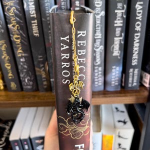 May include: A black and gold dragon bookmark with a chain. The bookmark is on a book titled "Fourth Wing" by Rebecca Yarros. The book is on a bookshelf with other books.