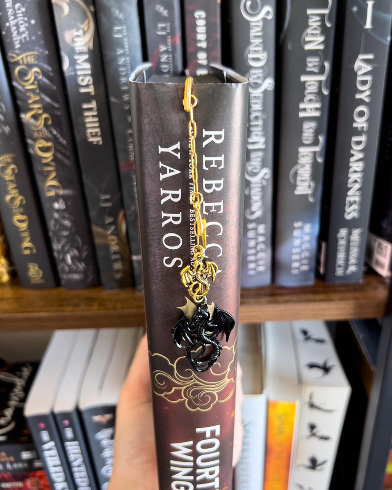 May include: A black and gold dragon bookmark with a chain. The bookmark is on a book titled "Fourth Wing" by Rebecca Yarros. The book is on a bookshelf with other books.