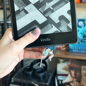 May include: A black Kindle e-reader with a silver bookmark charm featuring a sword, a butterfly, and a fox. The bookmark is attached to the bottom of the e-reader.