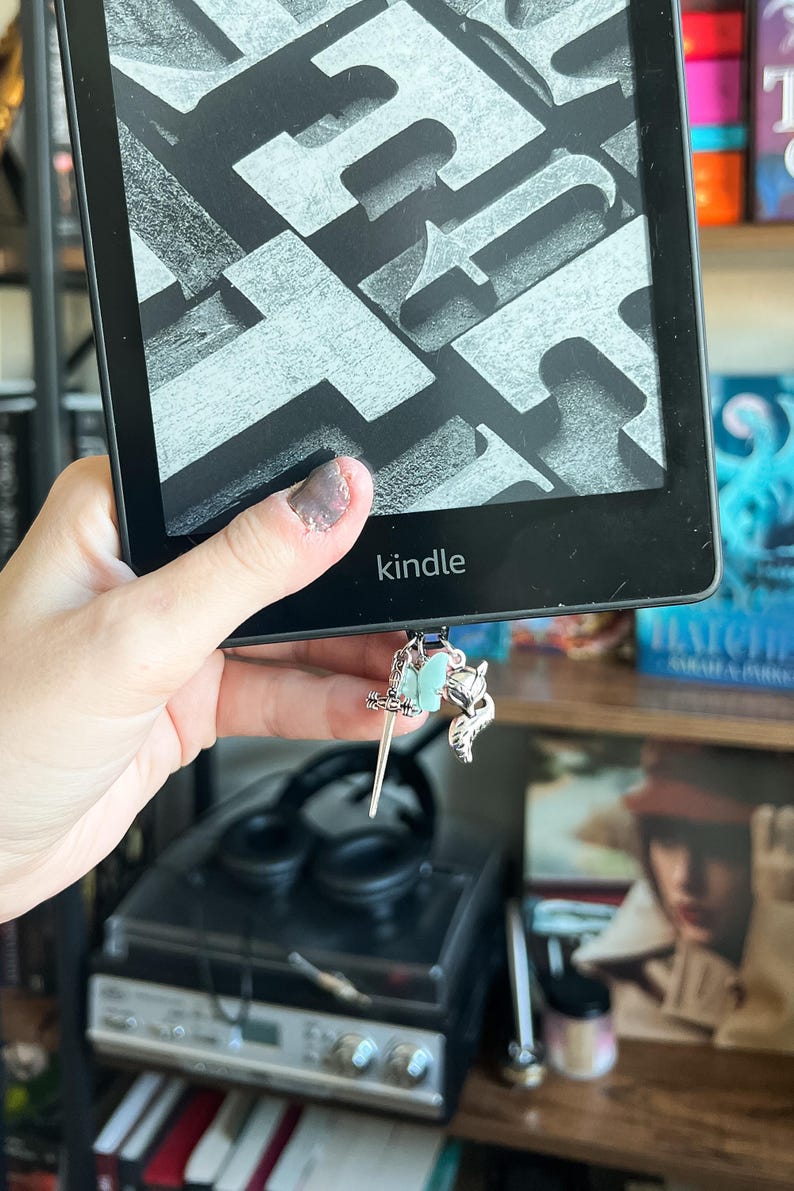 May include: A black Kindle e-reader with a silver bookmark charm featuring a sword, a butterfly, and a fox. The bookmark is attached to the bottom of the e-reader.