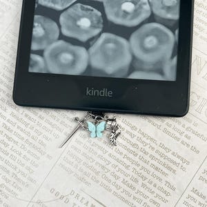 May include: A silver bookmark with a blue butterfly charm and a sword charm. The bookmark is attached to a Kindle e-reader. The text on the page reads "Dream of the Good Things to Come".