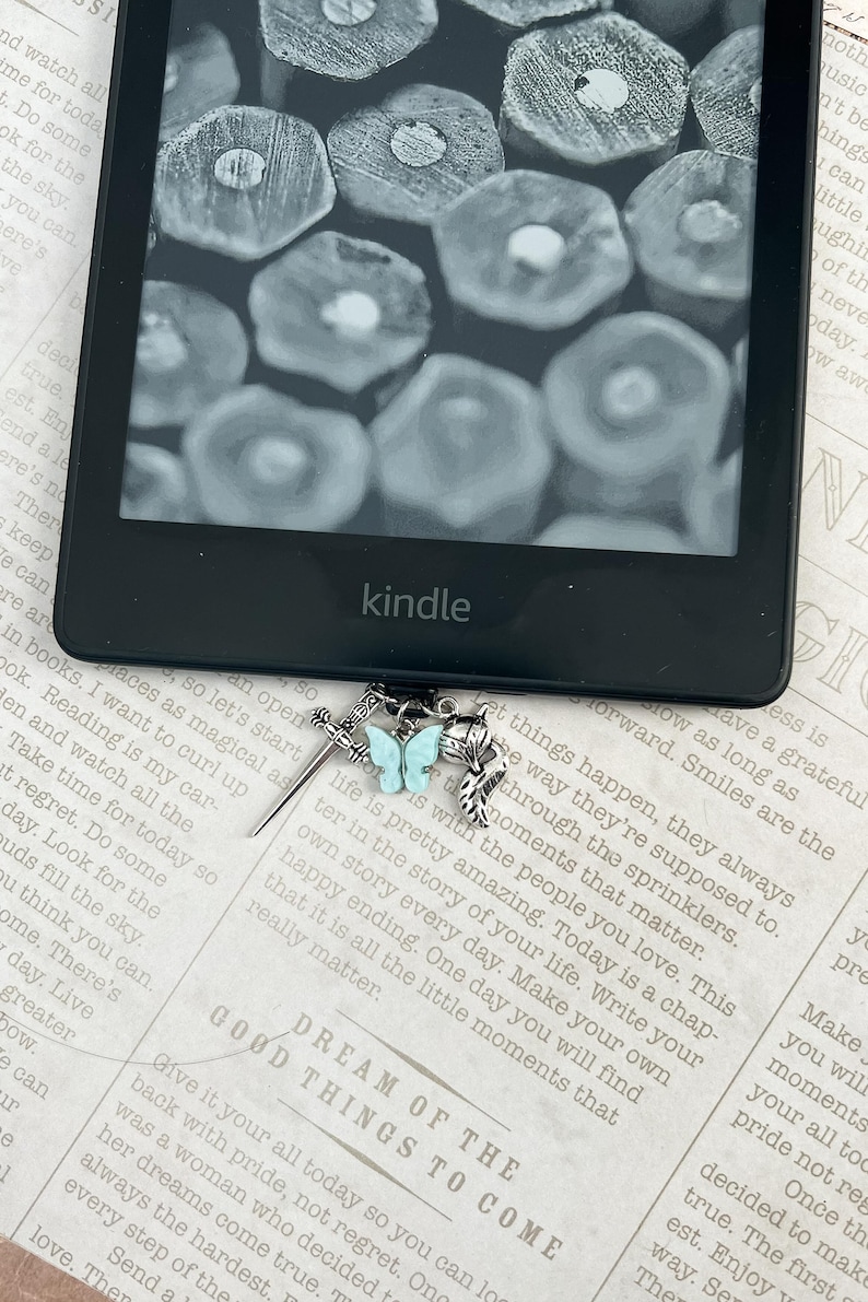 May include: A silver bookmark with a blue butterfly charm and a sword charm. The bookmark is attached to a Kindle e-reader. The text on the page reads "Dream of the Good Things to Come".