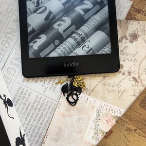 May include: A black and gold dragon bookmark charm with a lightning bolt detail. The charm is attached to a black e-reader with the word "kindle" printed on the front. The e-reader is lying on a surface covered in vintage paper with handwritten text.