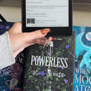Puede incluir: Primer plano de un libro de bolsillo titulado "Powerless" de Lauren Roberts. La portada del libro presenta una espada con una corona en la parte superior, rodeada de hojas verdes y flores moradas. El libro forma parte de una serie llamada "The Powerless Trilogy".