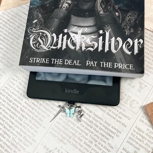 May include: A silver bookmark with a sword, a feather, and a butterfly charm. The butterfly is light blue and the bookmark is hanging from a Kindle e-reader. The text on the Kindle screen reads "Good things to come. Dream of the..."