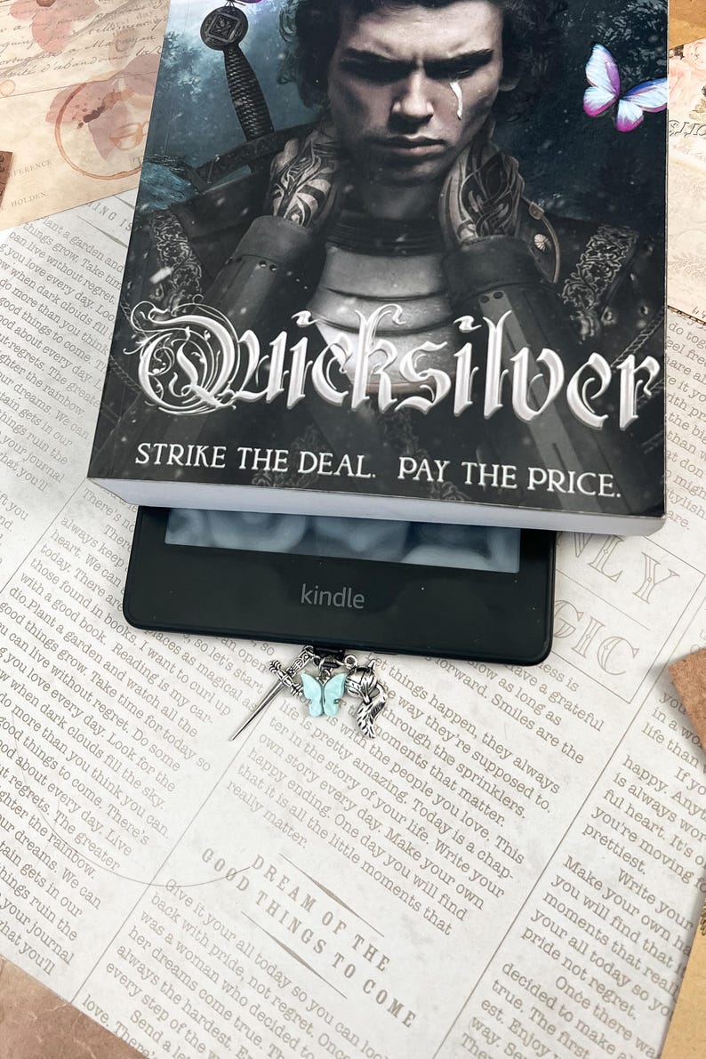 May include: A silver bookmark with a sword, a feather, and a butterfly charm. The butterfly is light blue and the bookmark is hanging from a Kindle e-reader. The text on the Kindle screen reads "Good things to come. Dream of the..."