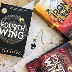 May include: Three fantasy novels with a dragon theme. The books are titled "Fourth Wing" and "Iron Flame". The books are on a table with a gold bookmark and a dragon charm.