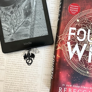 May include: A black Kindle e-reader with a black dragon bookmark attached. The Kindle is displaying a black and white image of a glass inkwell and a pen. The book cover is red with gold lettering and the text "FOURTH" and "REBECCA" in white.