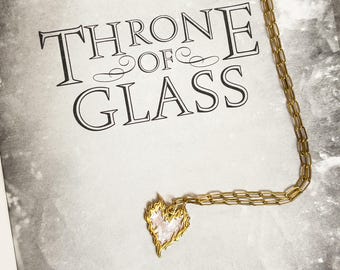 May include: Gold-colored necklace with a heart-shaped pendant. The pendant has a flame-like design around a white center. The necklace is on a book with the words "THRONE OF GLASS" in black.