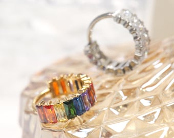 Puede incluir: Dos anillos de oro con piedras preciosas de colores. El primer anillo tiene un arcoíris de piedras preciosas rectangulares. El segundo anillo tiene piedras preciosas redondas transparentes.